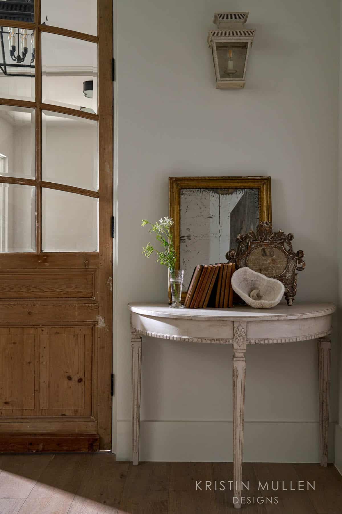 Demilune console table with antique mirror, stacked books, and decorative objects beside an open pine paneled door