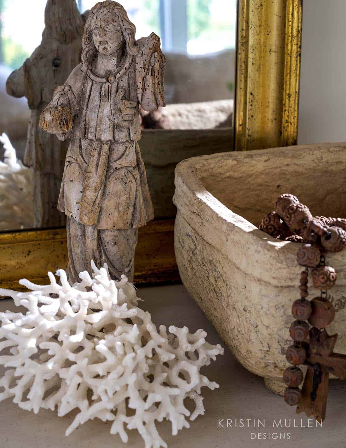 Close-up vignette of a weathered wooden angel figurine, coral piece, and stone vessel with rosary beads on a shelf