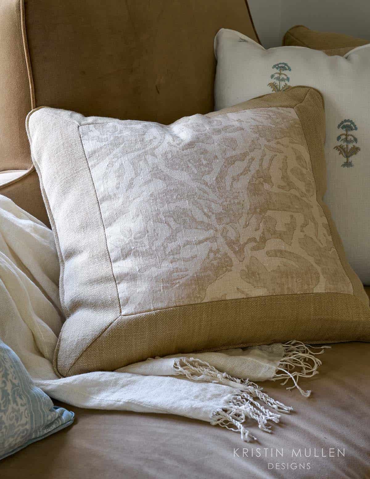 Textured linen throw pillow with subtle floral pattern on a tan velvet sofa, layered with an embroidered pillow and fringed throw