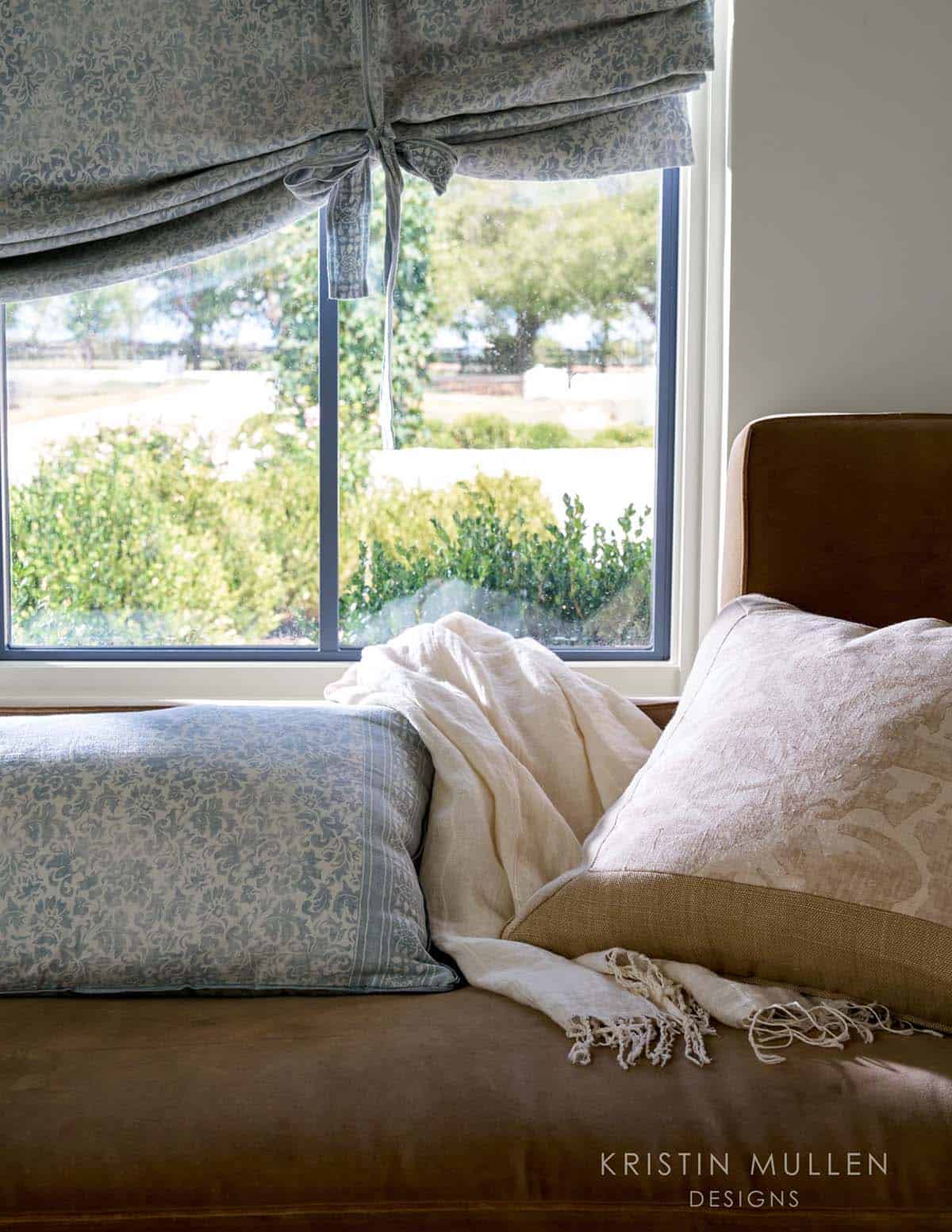 Corner window seat with blue floral bolster, ivory linen throw, and patterned pillows overlooking a garden view