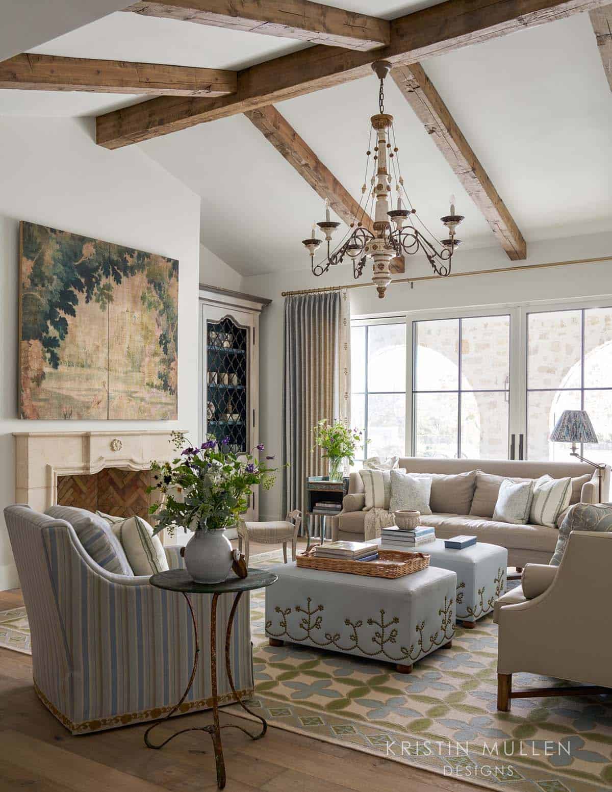 Living room with vaulted wood-beamed ceiling, ornate chandelier, stone fireplace, and blue-and-cream striped armchairs