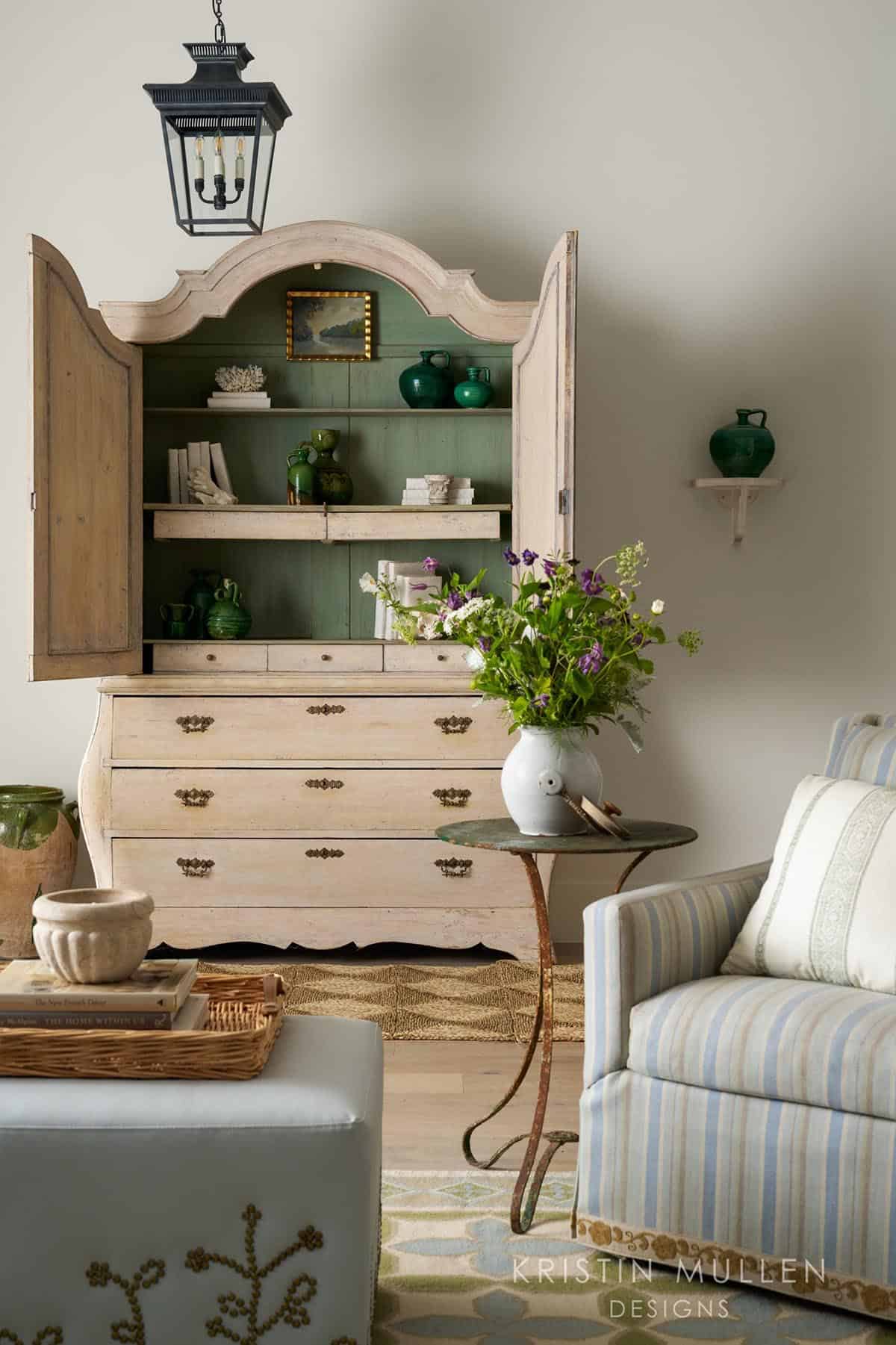 Open antique armoire with sage-green interior displaying green glass vessels, books, and decorative objects beside a striped armchair