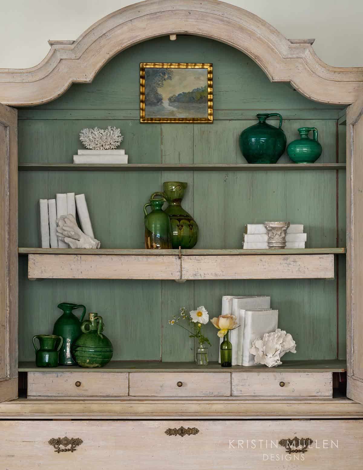 Close-up of an armoire's sage-green shelving filled with green glass jugs, framed artwork, books, and coral decor