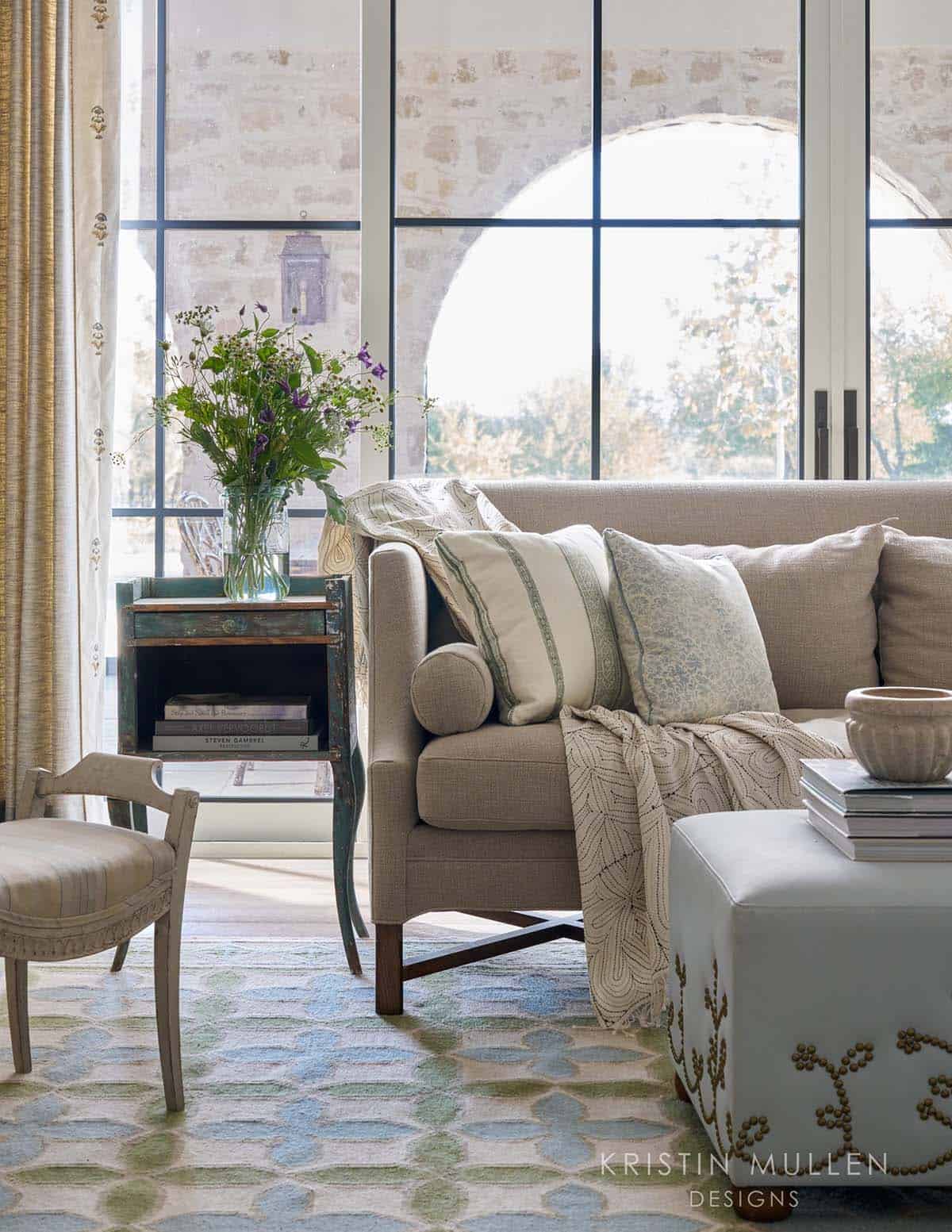 Neutral sofa with striped and floral pillows beside a distressed blue side table, arched windows showing stone exterior