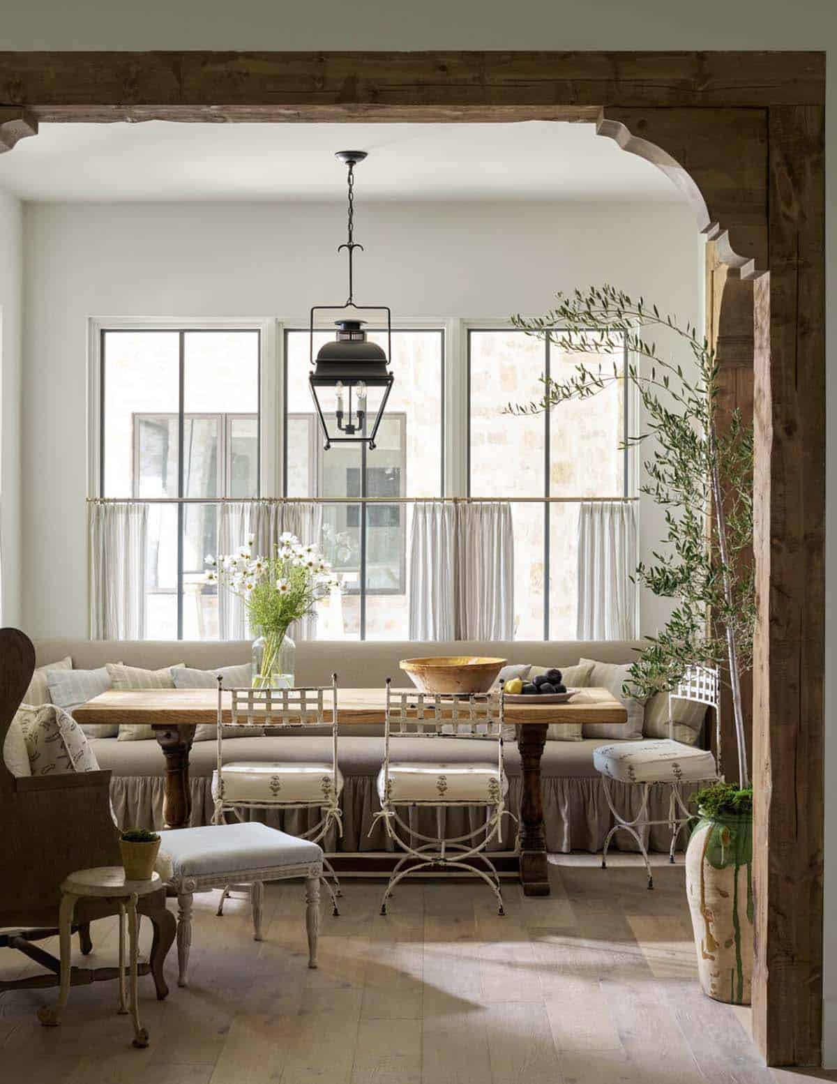 Breakfast nook with wood beams, rustic trestle table, iron chairs, upholstered banquette, olive tree, and lantern pendant