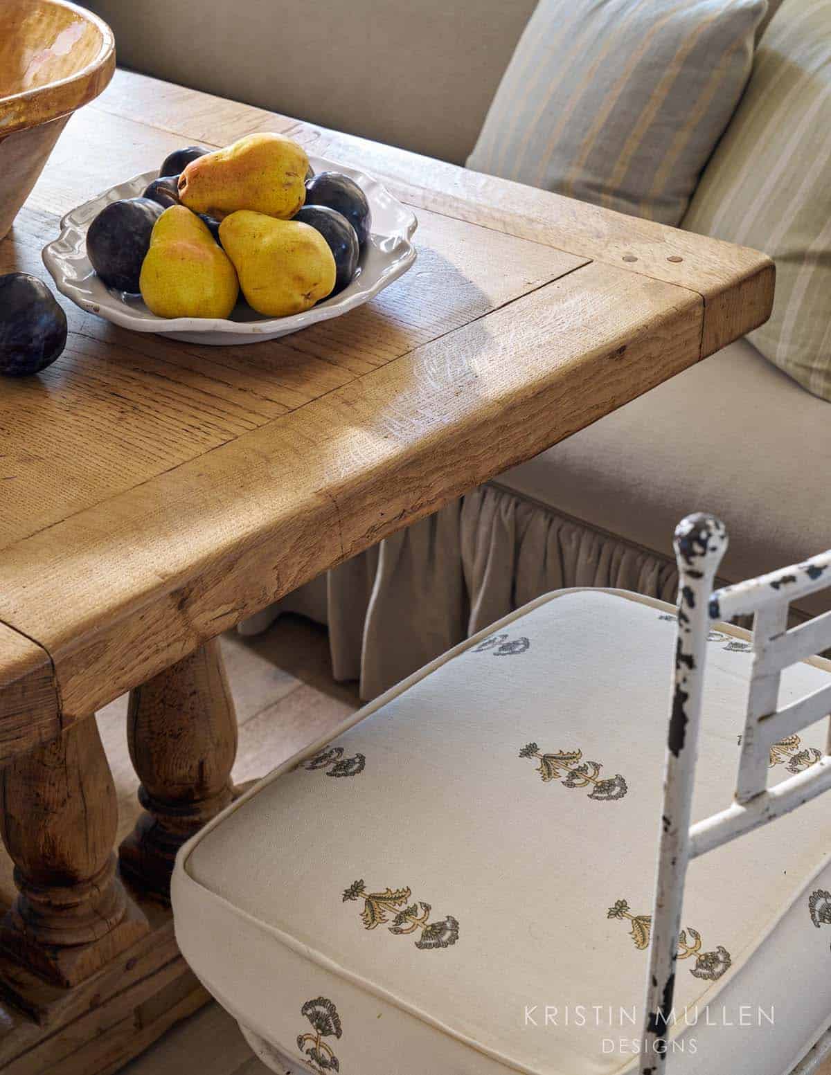 Close-up of a rustic wood table edge with a bowl of pears and plums beside an antique iron chair with block-print cushion