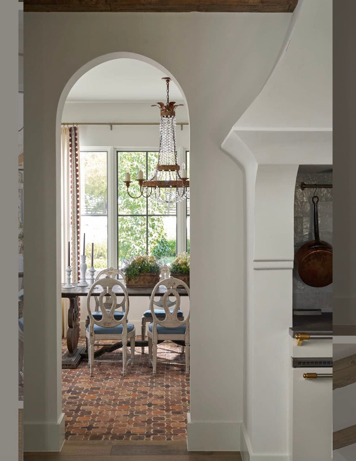 View through an arched doorway into a dining room with brick floor, beaded chandelier, and Swedish-style dining chairs