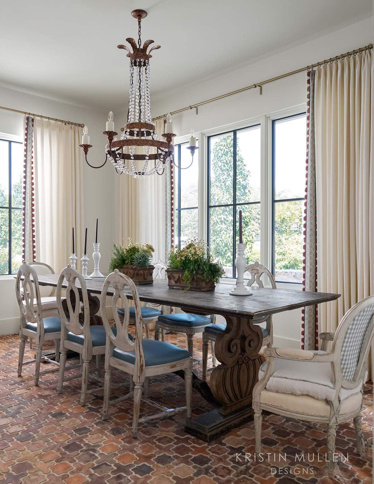 Dining room with beaded iron chandelier, brick floor, cream drapes with red trim, and painted French chairs with blue seats