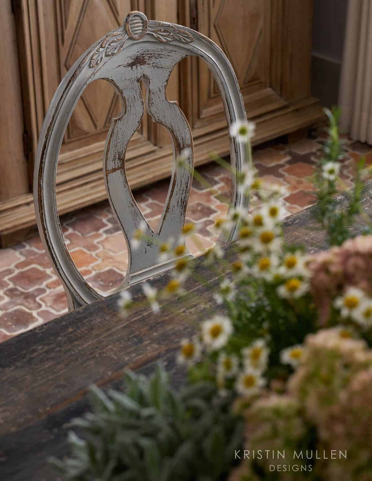 Close-up of a distressed white painted dining chair back with daisies and hydrangeas in the foreground, brick floor behind