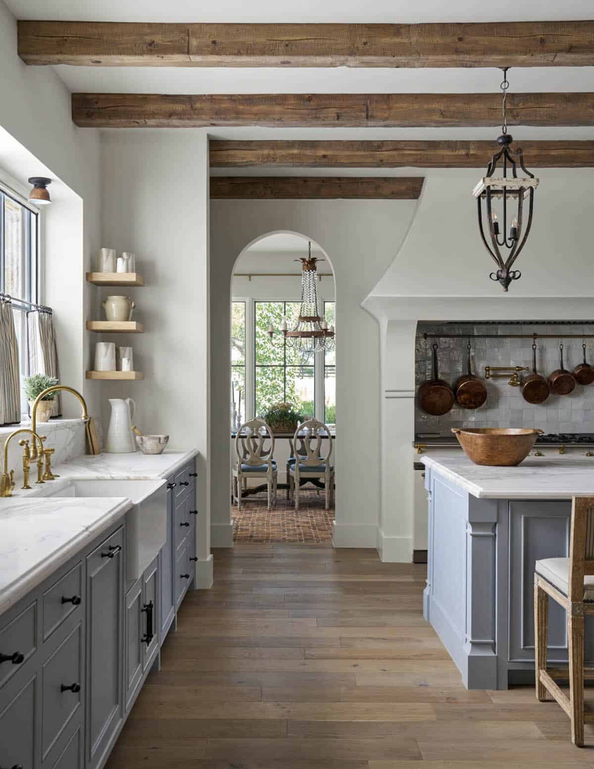 Kitchen with wood beams, gray cabinetry, brass fixtures, copper pots, and a view through an arch to the dining room