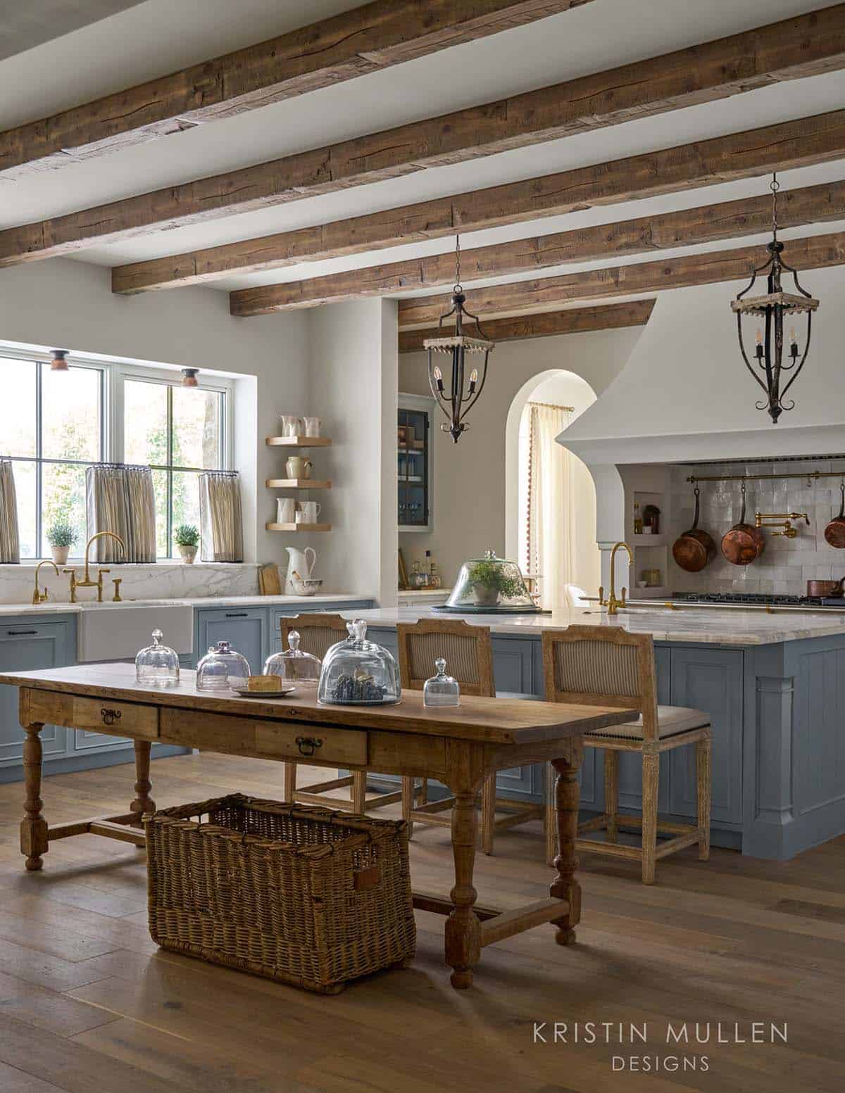 Kitchen with wood beams, blue-gray cabinets, brass fittings, hanging copper pots, and a rustic wood table with glass cloches