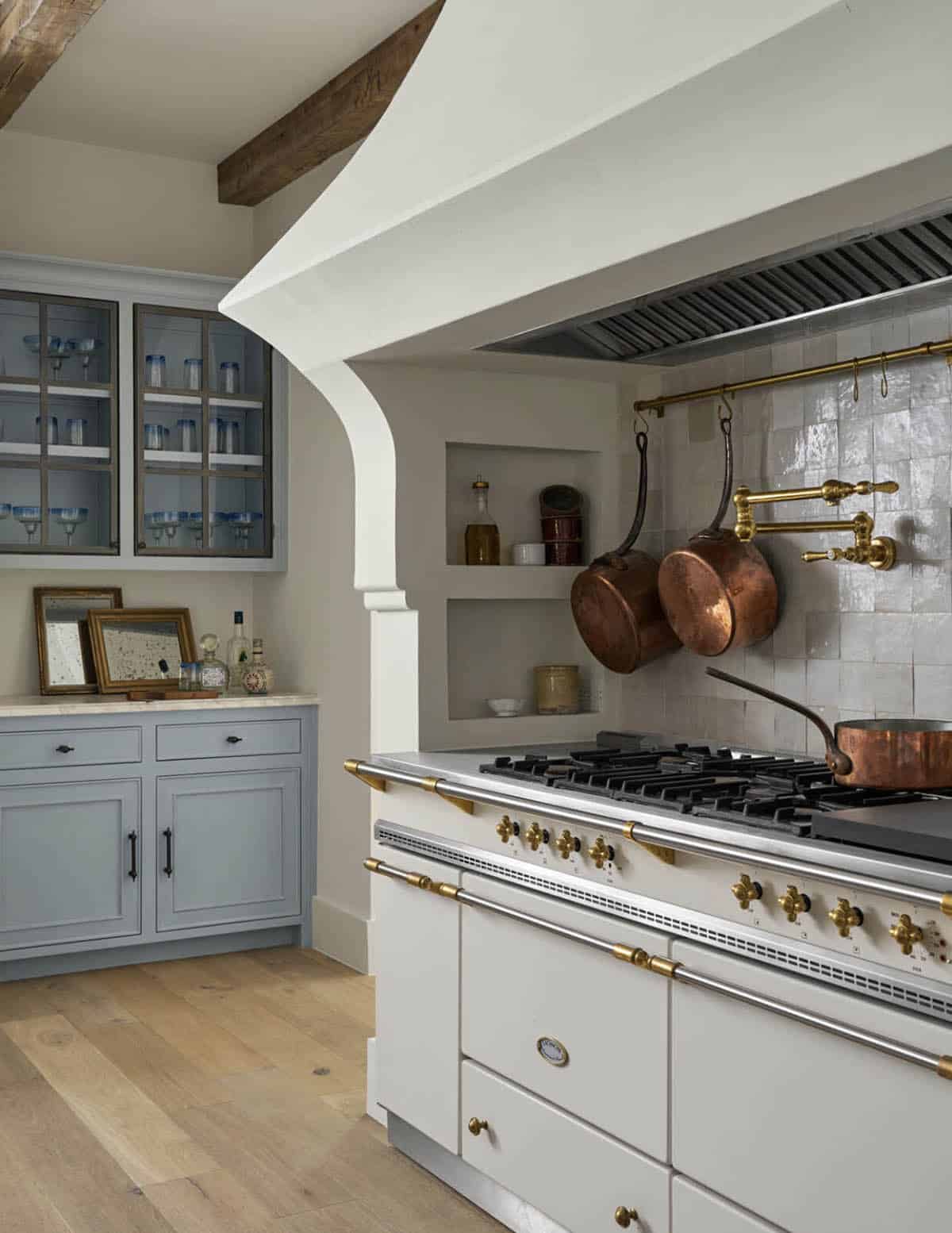 Range and hood detail with hanging copper pots, brass pot filler, zellige tile backsplash, and blue glass-front cabinet