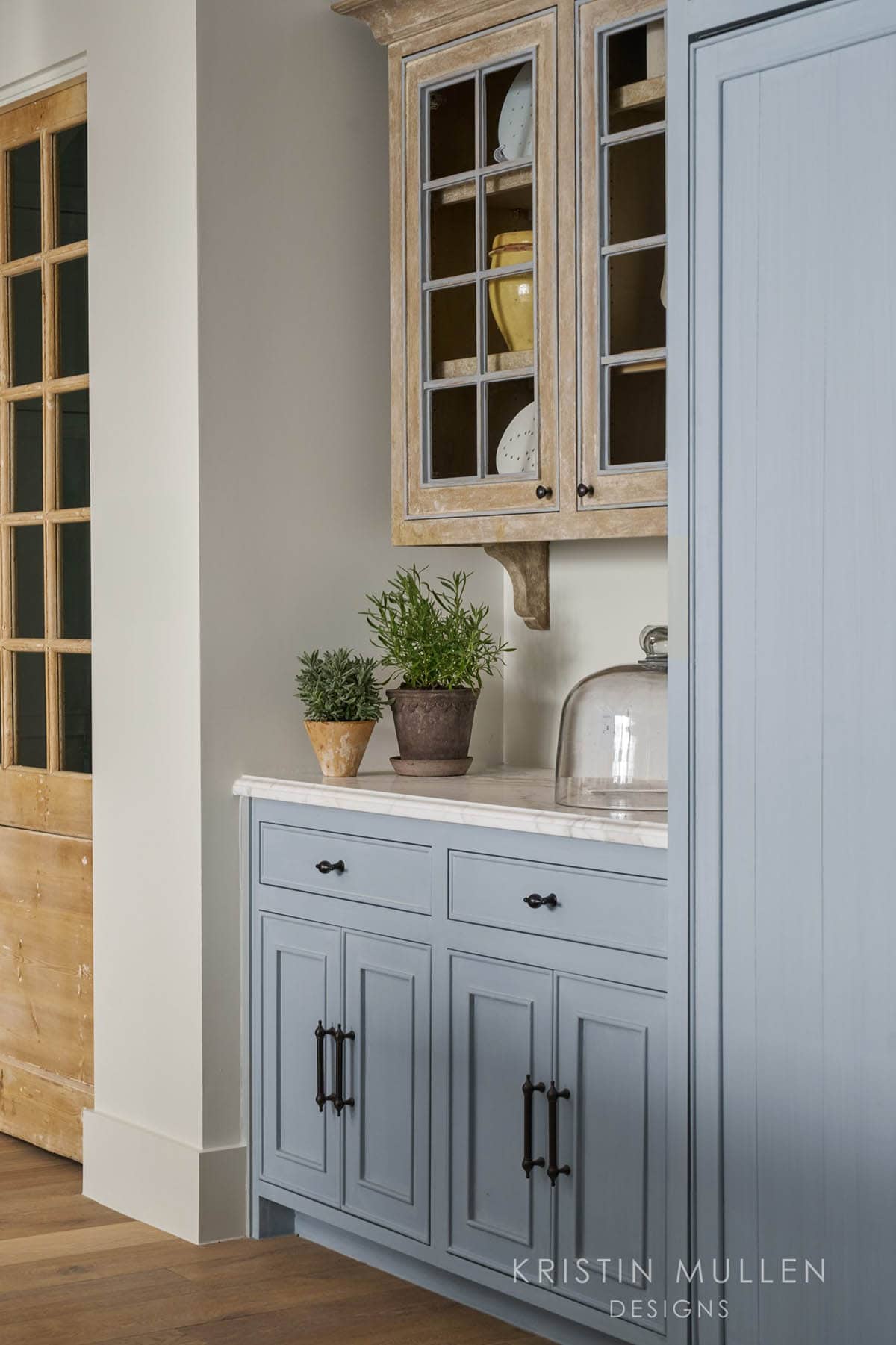 Kitchen cabinetry detail with weathered wood glass-front cabinet, blue-gray drawers, brass hardware, and potted herbs