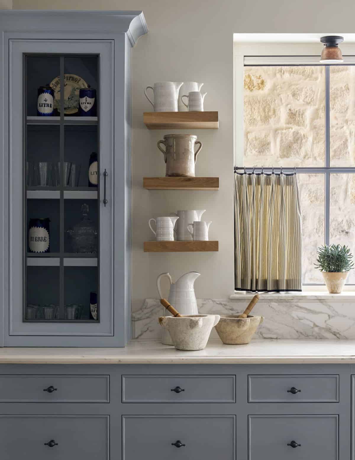 Kitchen shelving with white ceramic pitchers, stoneware crocks, marble counter, and a striped café curtain at the window