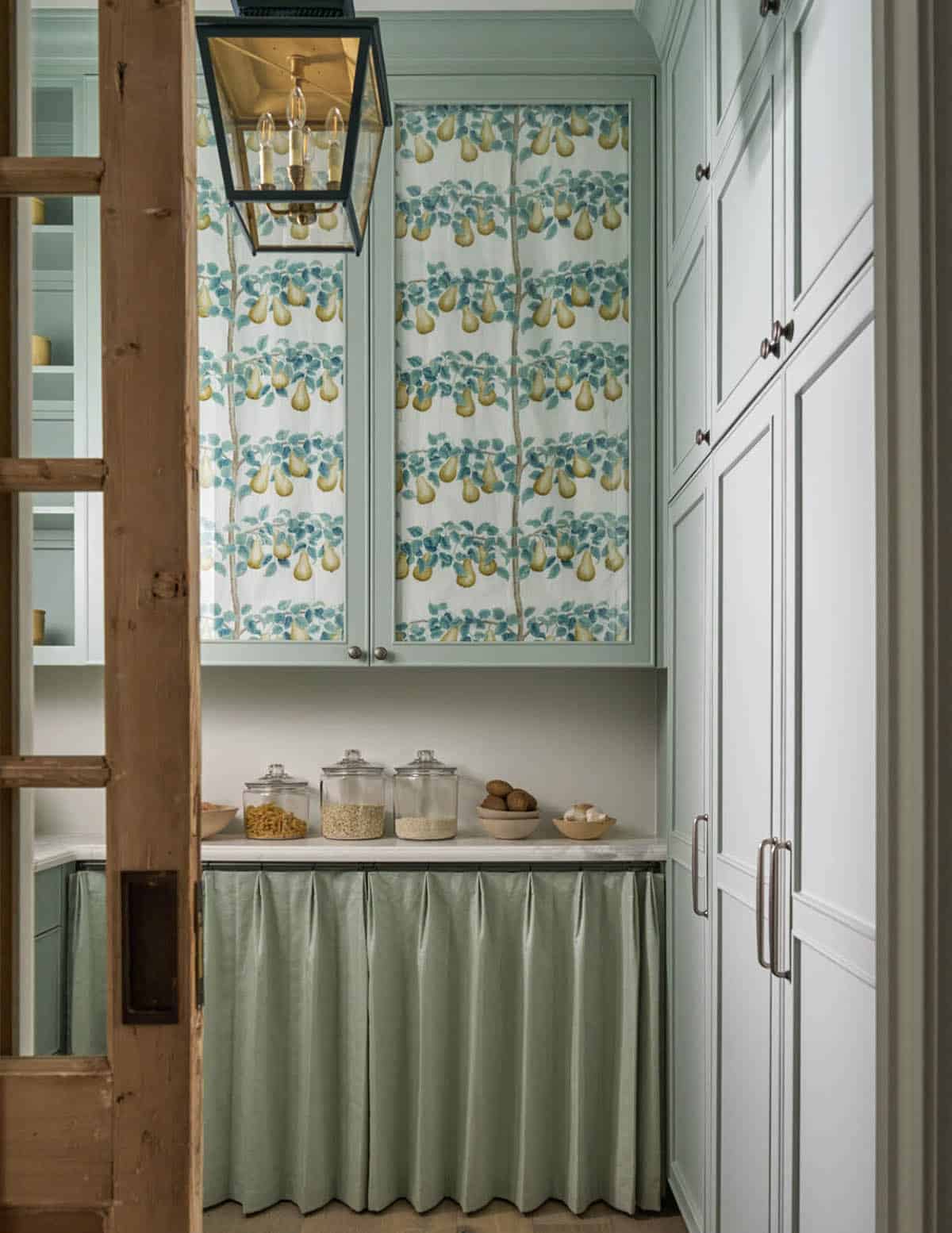 Butler's pantry with sage-green cabinetry, pear-print fabric panels, lantern pendant, and glass storage jars