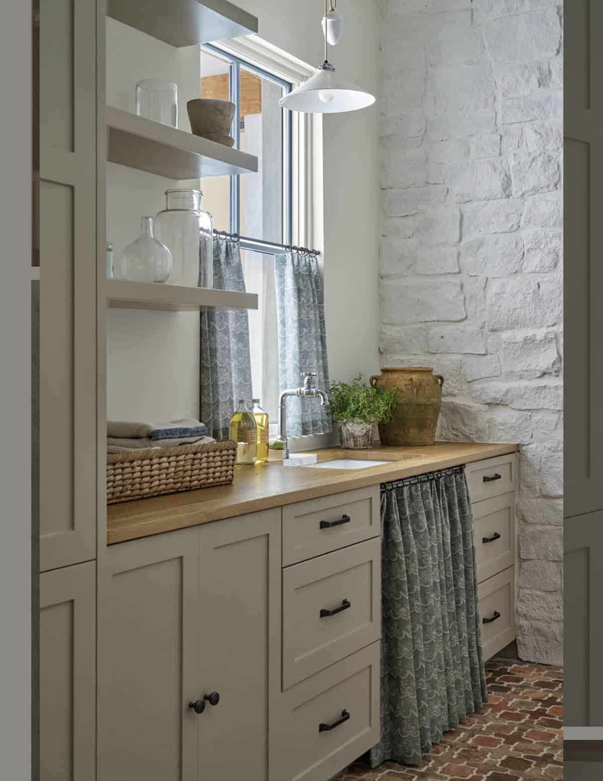 Utility sink area with open shelving, wood countertop, patterned curtain skirt, and whitewashed stone wall