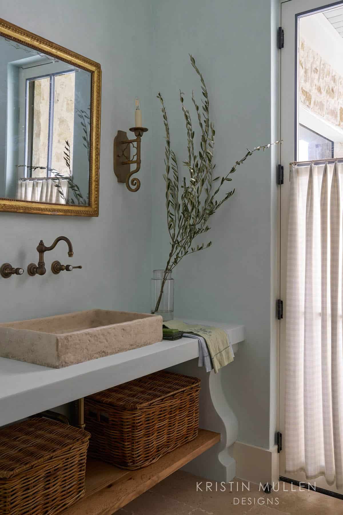 Powder room vanity with carved stone vessel sink, brass wall-mounted faucet, gilt mirror, and wicker baskets below