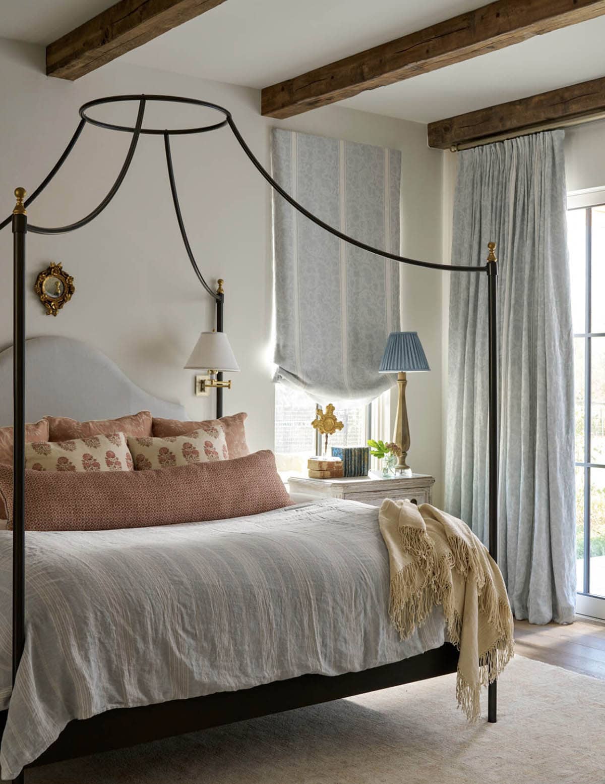 Bedroom with black iron canopy bed, blush and cream pillows, wood beams, and blue floral striped roman shade