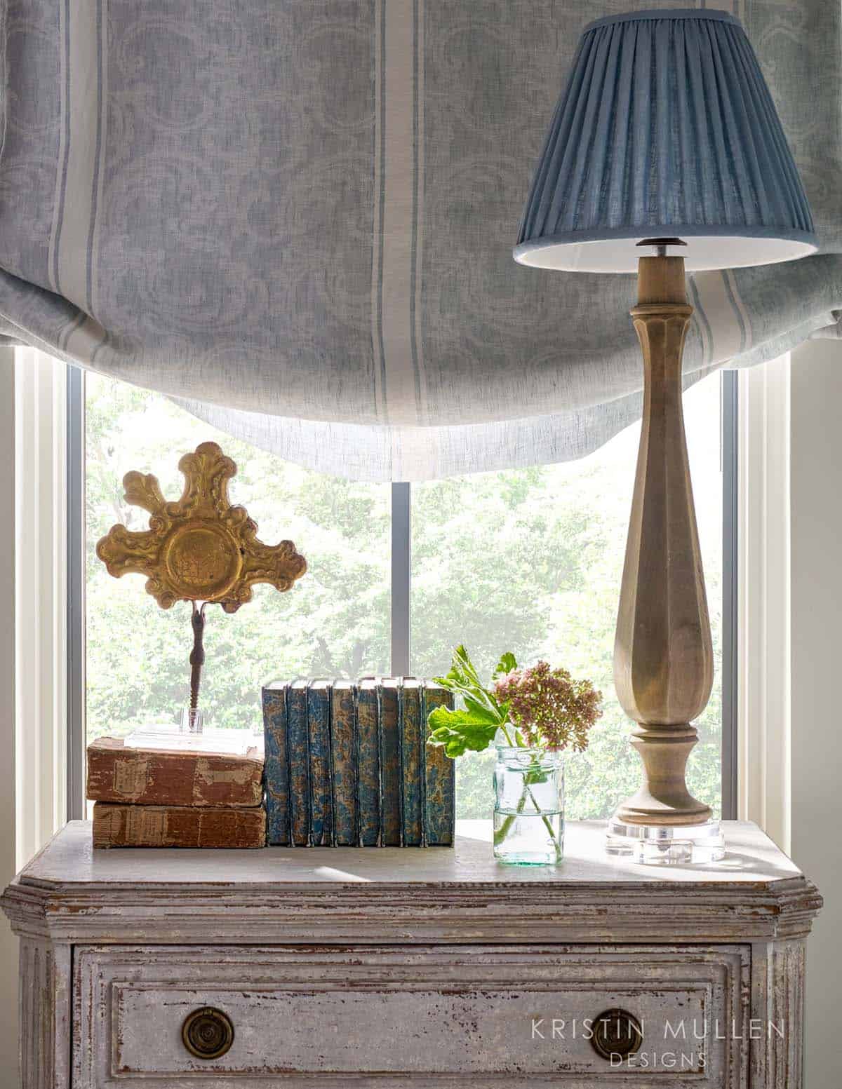 Bedside vignette with antique books, brass ornament, wood table lamp with blue pleated shade, and striped roman shade