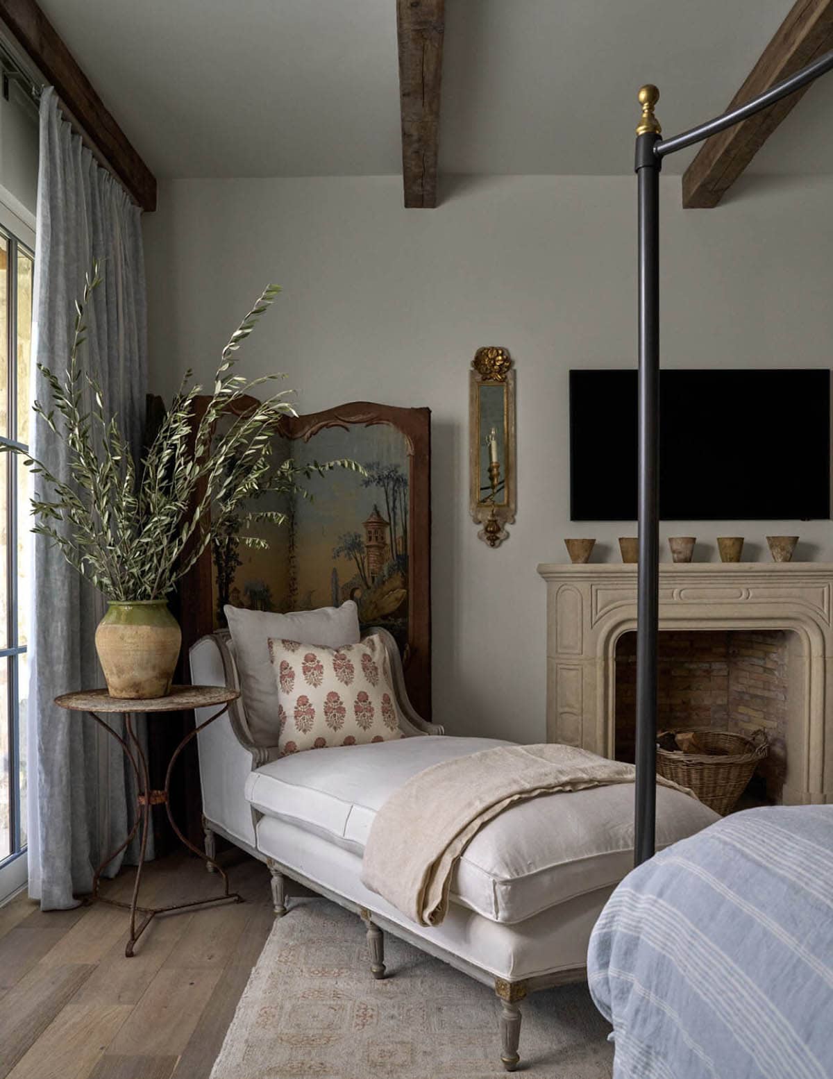 Bedroom seating area with upholstered chaise lounge, painted screen, stone fireplace, and mounted television