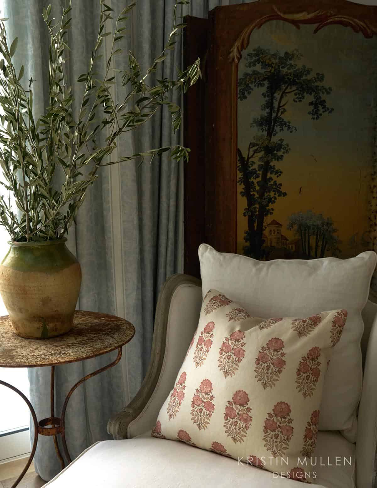 Close-up of a chaise lounge with pink floral pillow, antique painted screen, and rustic olive branches in a ceramic urn