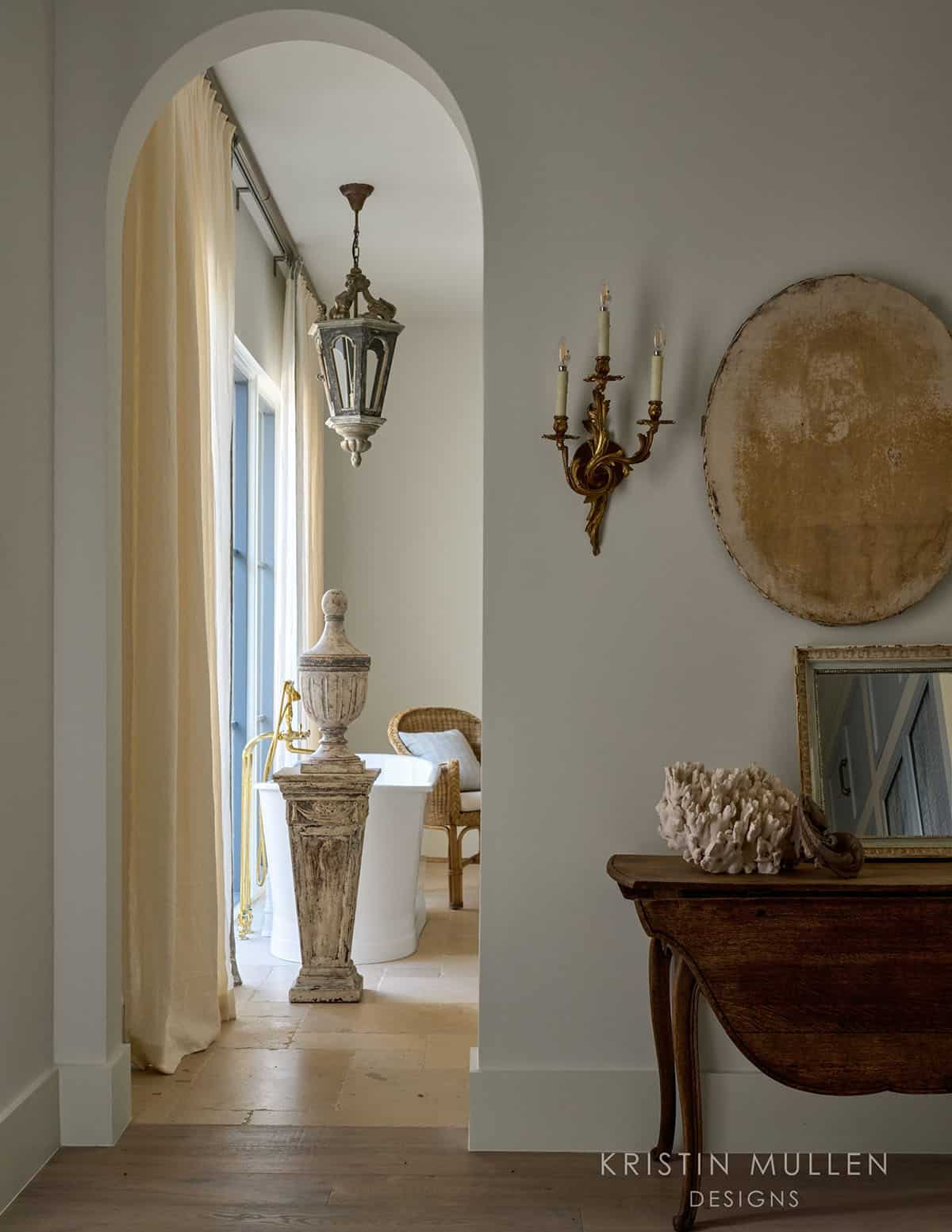 View through an arched doorway into a bathroom with freestanding tub, wicker chair, and antique stone urn on pedestal