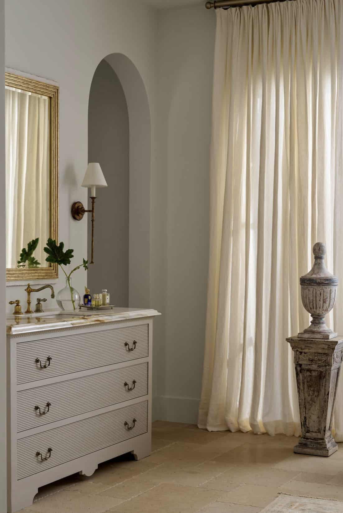 Gray dresser-style vanity with brass fixtures beside cream drapery and a weathered stone urn on a pedestal