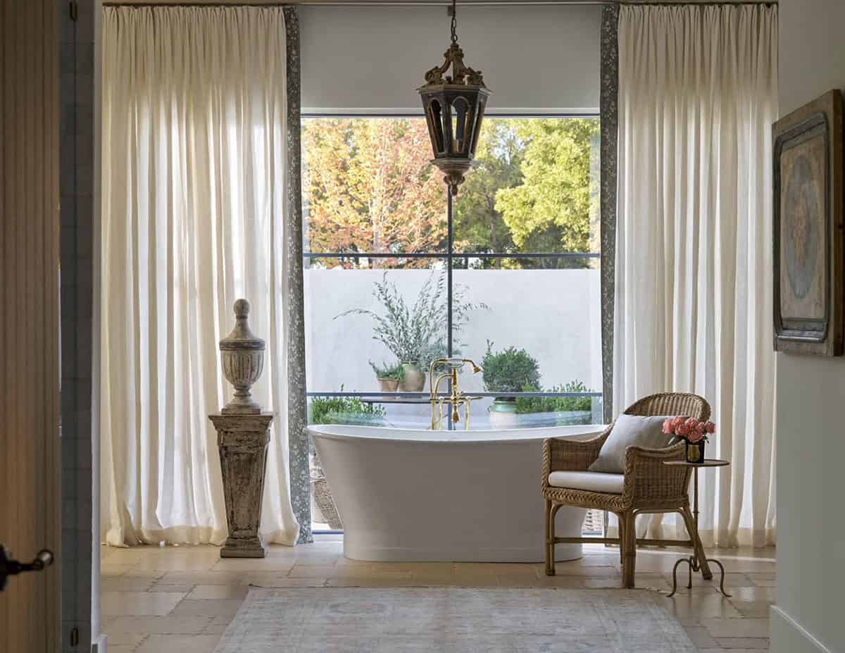 Freestanding soaking tub beneath a large window with brass fixtures, wicker armchair, and antique iron lantern above