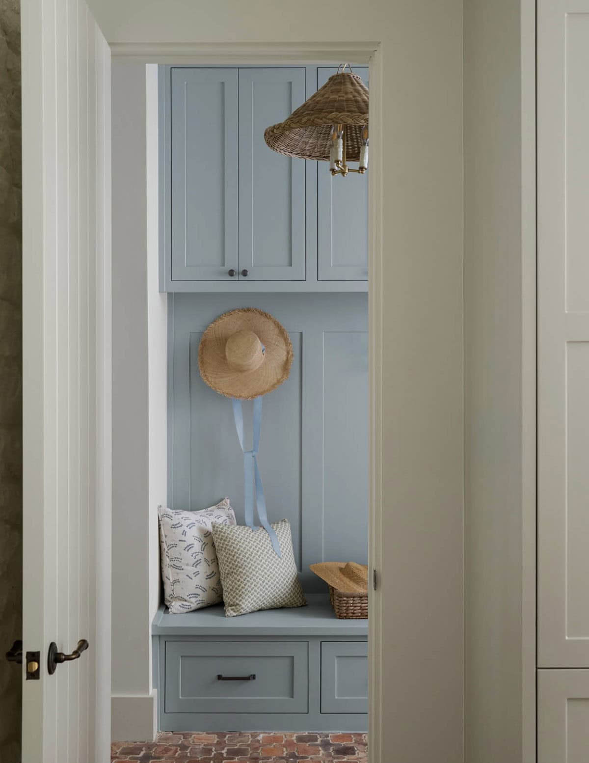 View into a mudroom nook with blue cabinetry, woven pendant light, straw sun hat, and patterned bench pillows