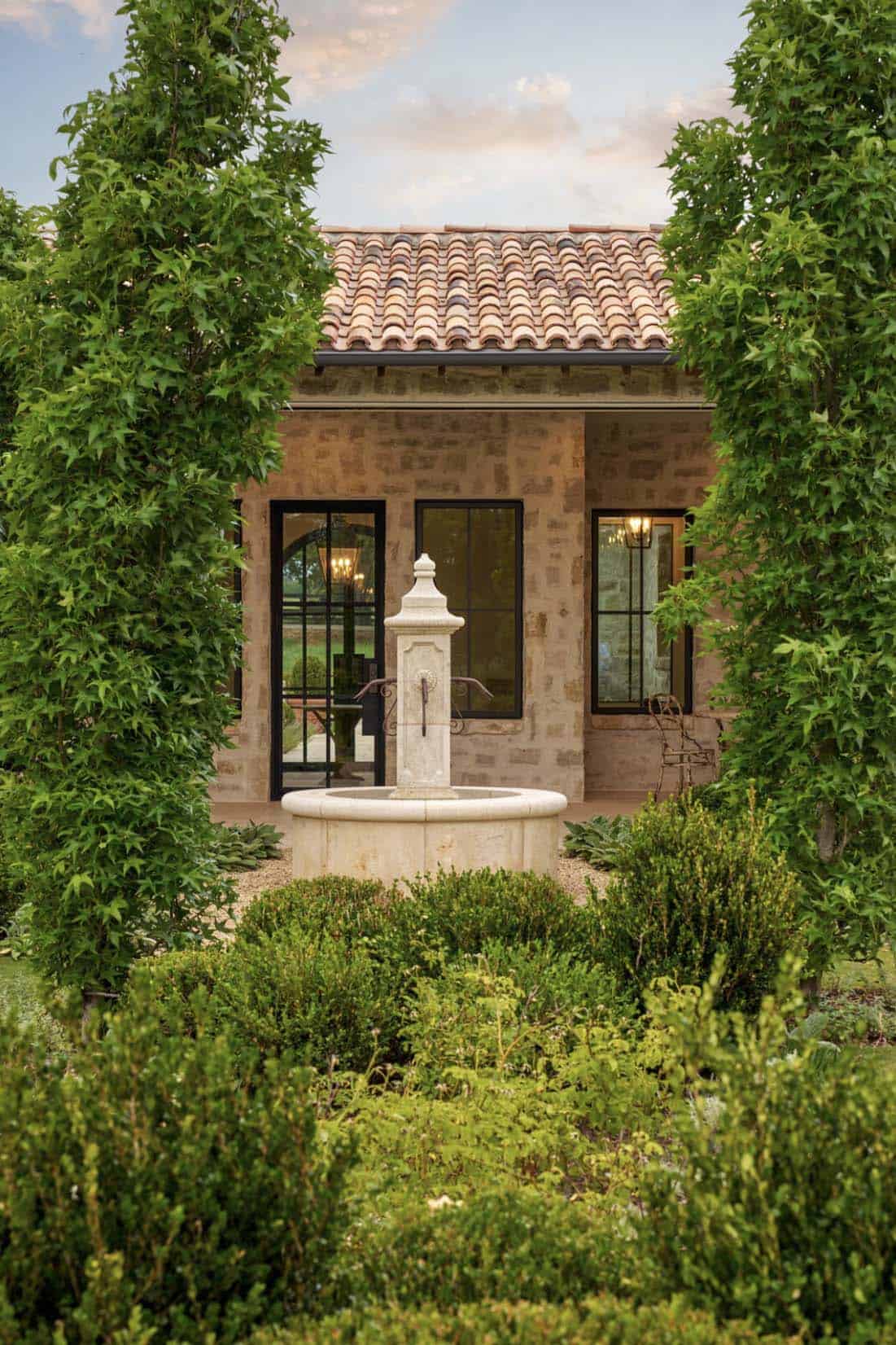 Mediterranean home exterior with terracotta tile roof and stone fountain, framed by tall cypress trees and boxwood hedges
