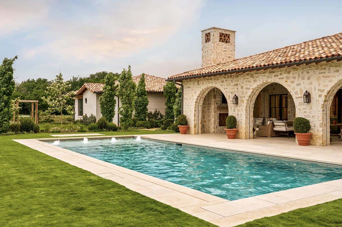 Rectangular swimming pool with water jets beside a stone Mediterranean home with arched covered patio and chimney