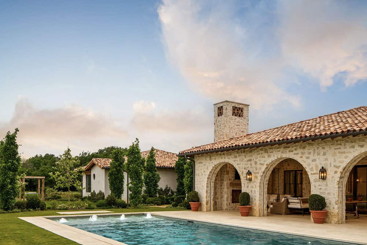 Wide view of stone Mediterranean home with arched loggia, tall chimney, pool, and dramatic cloud-filled sky