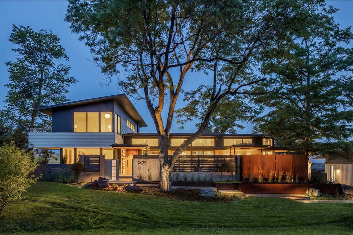 Modern two-story home with dark cladding lit up at dusk, surrounded by mature trees and a landscaped front yard