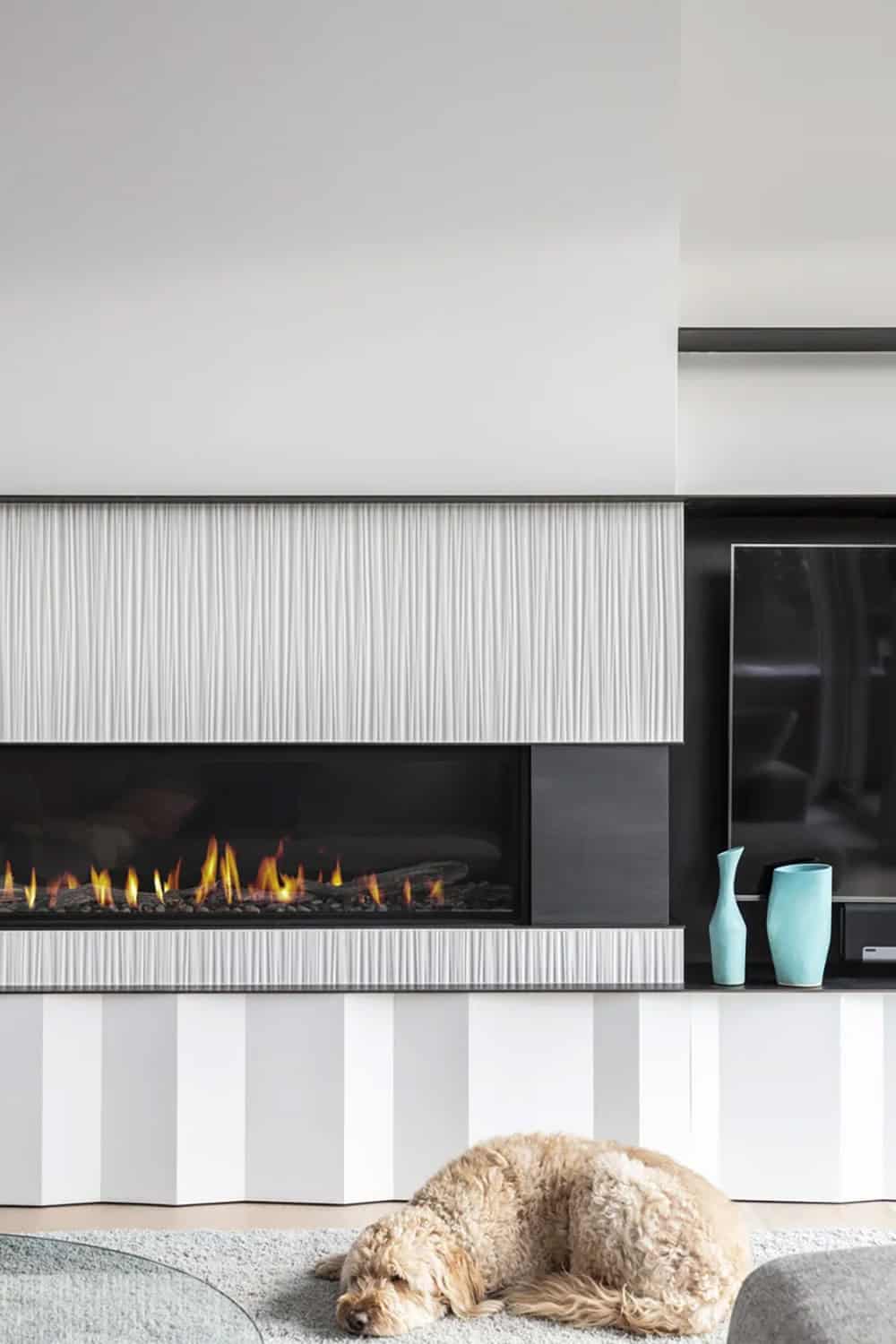 Linear gas fireplace with ribbed white paneling, teal vases, and a shaggy dog napping on the rug below