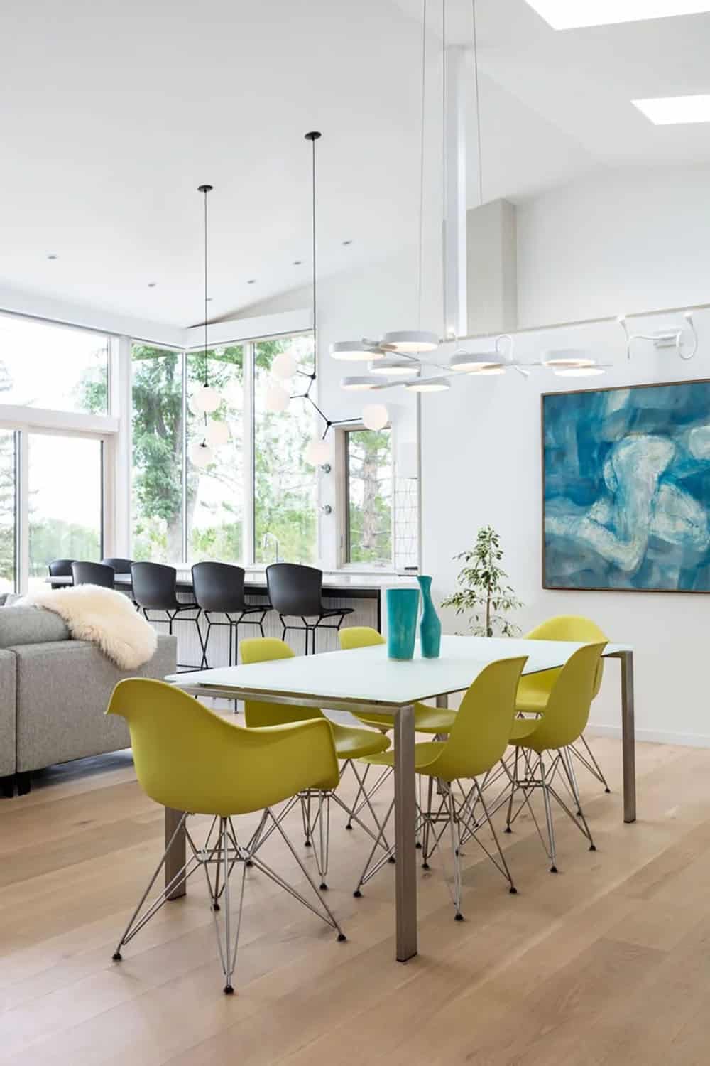 Dining area with yellow chairs and glass table beneath a modern chandelier, blue abstract painting on the wall