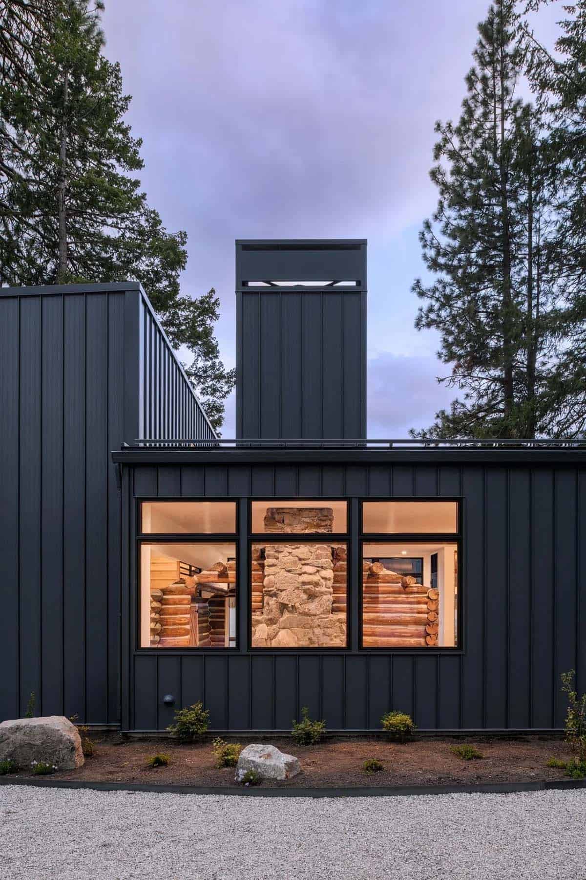 Close-up of black metal-clad cabin exterior with windows revealing interior log walls and stone chimney