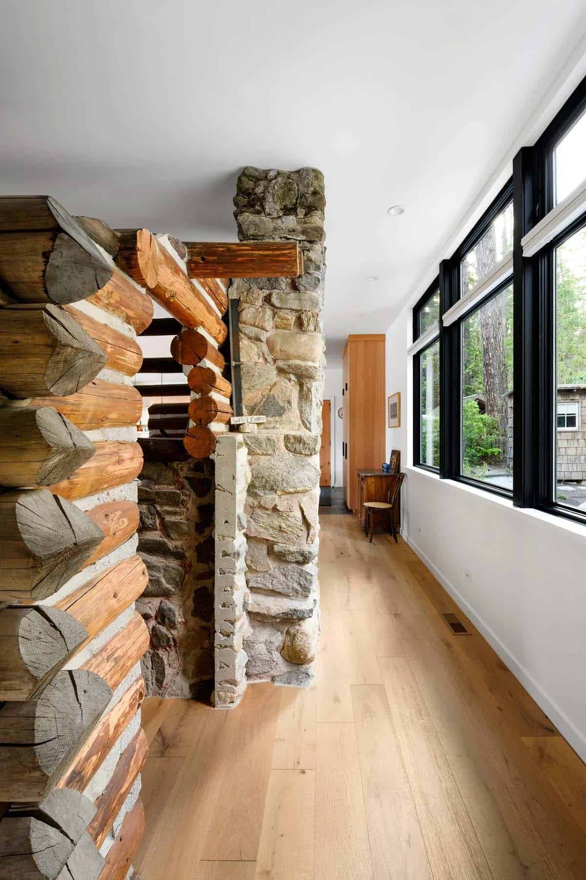 Hallway view past log cabin corner and stone chimney toward large black-framed windows