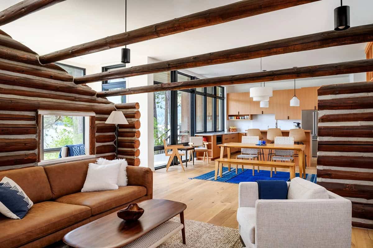 Living room framed by log cabin walls and exposed log ceiling beams, opening to kitchen and dining area
