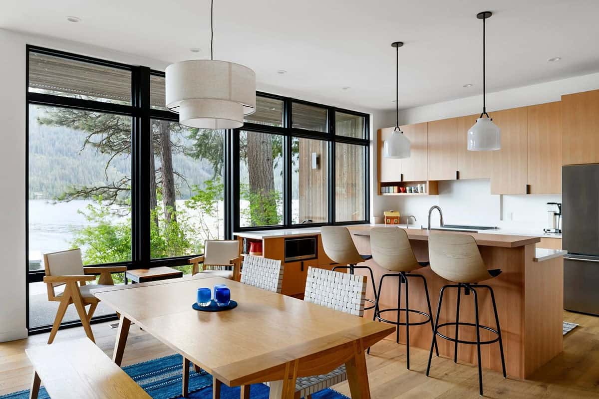 Dining area with wooden table and bar stools beside floor-to-ceiling windows overlooking the lake