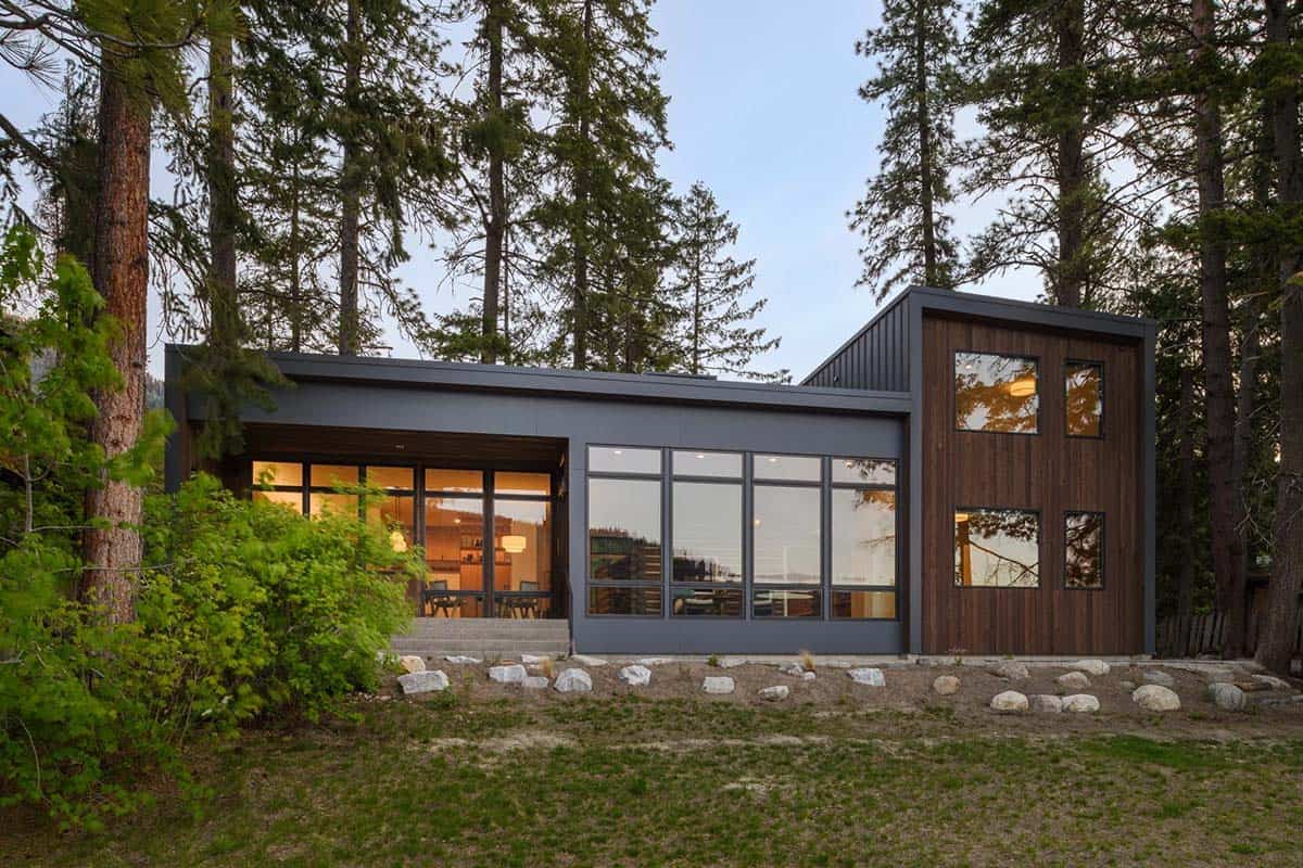 Twilight exterior of modern cabin with glowing windows, dark wood siding, and surrounding pine forest