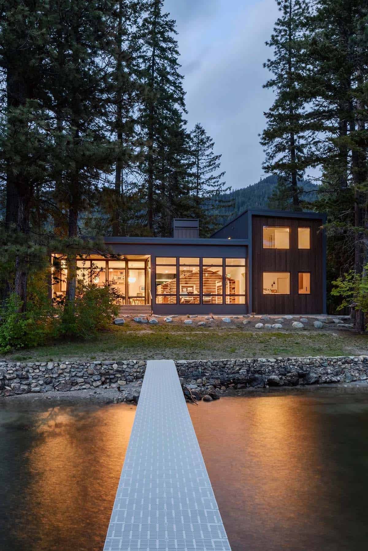 Dusk view of lakefront cabin from a dock, with warm interior lighting reflecting on the water
