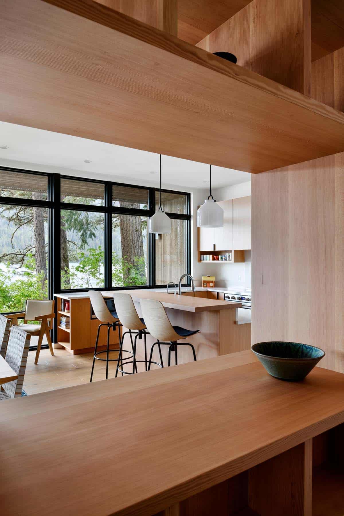 View through wood beams toward a bright kitchen with island seating and lake-facing windows
