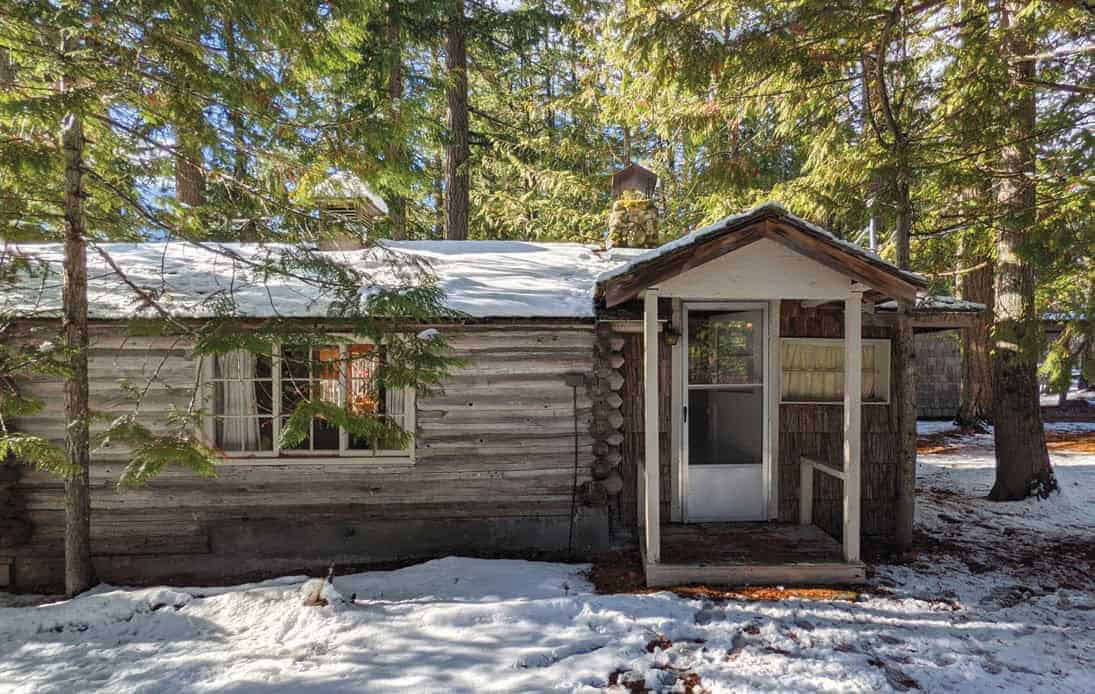 Original weathered log cabin exterior in snow, with covered entry porch and stone chimney