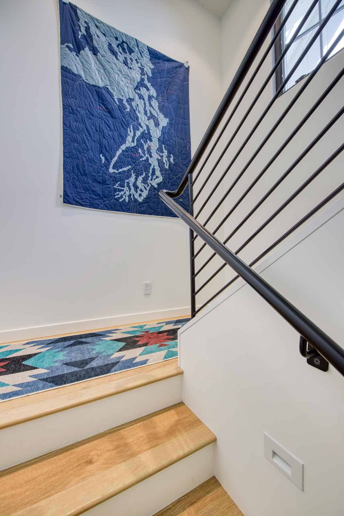 Staircase detail with black metal railing, patterned runner rug, and framed quilted map art