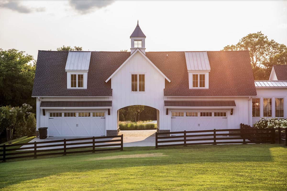 Garage building with white carriage doors, cupola, and black fencing at sunset