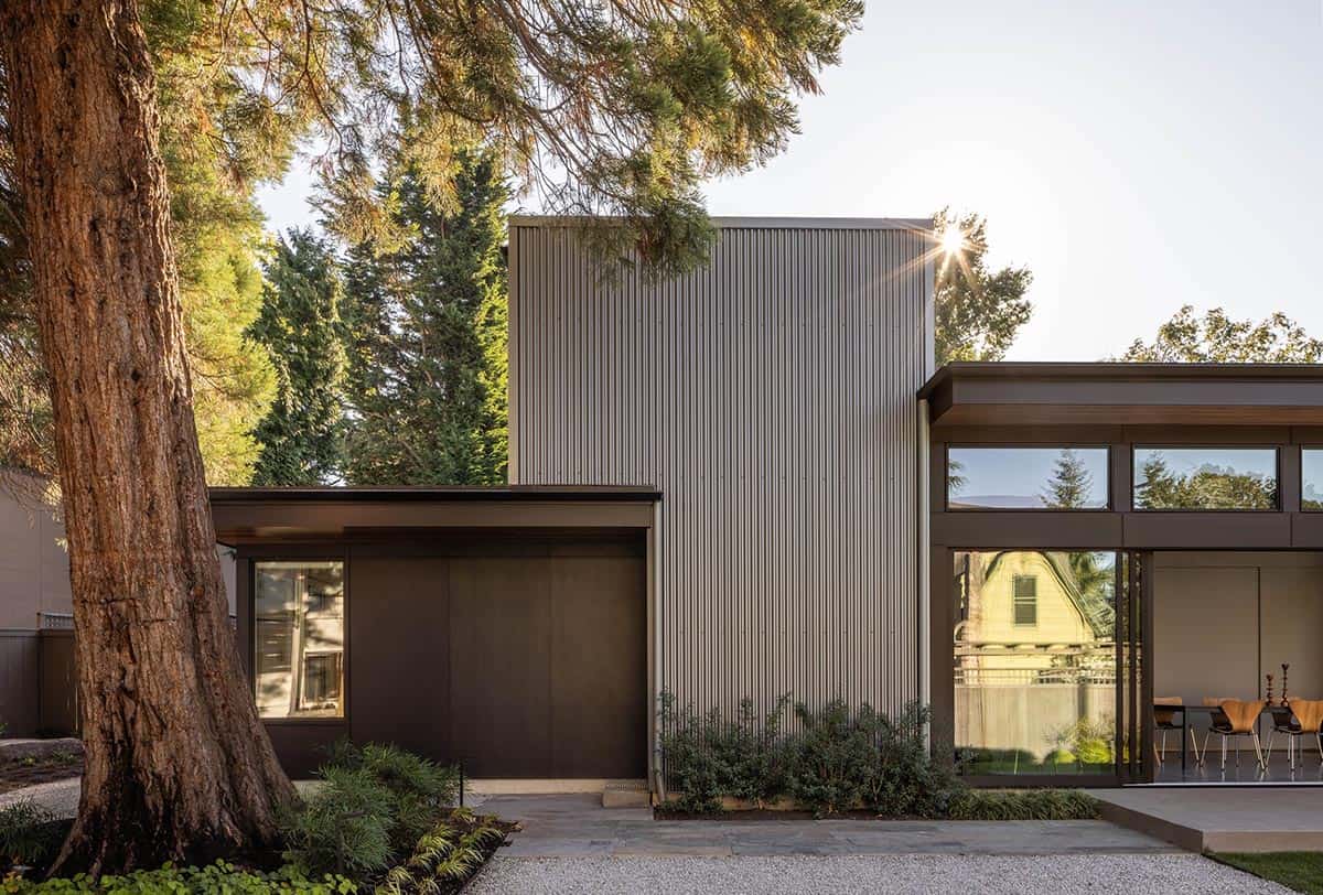 Corrugated metal and wood-clad exterior of the Quincho beside a sequoia trunk, sun flaring through branches