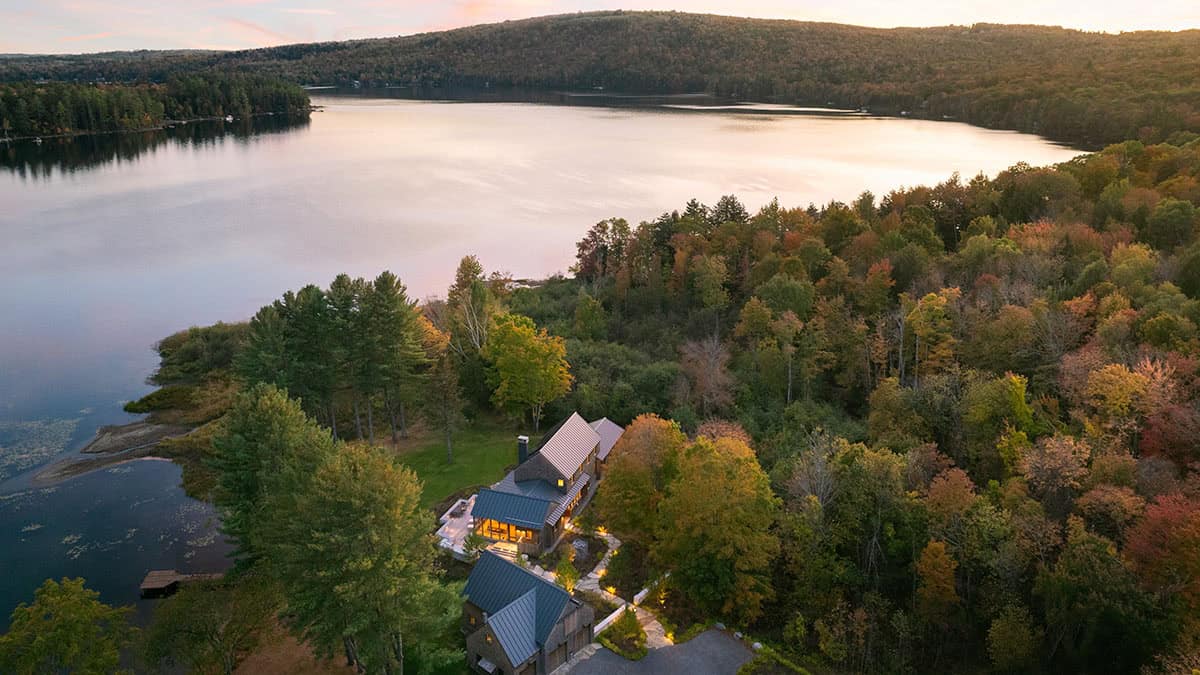 Aerial view of a modern lake house nestled among trees at sunset, with a large lake and forested hills in the background