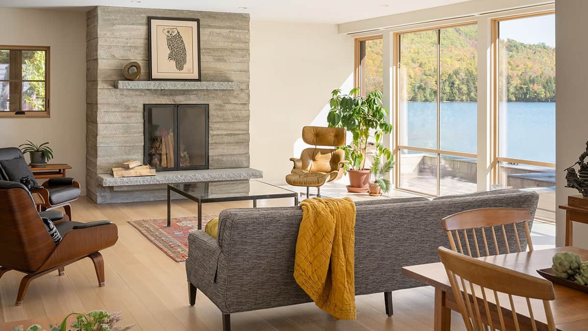 Living room with stone fireplace, mid-century chairs, and floor-to-ceiling windows facing the lake