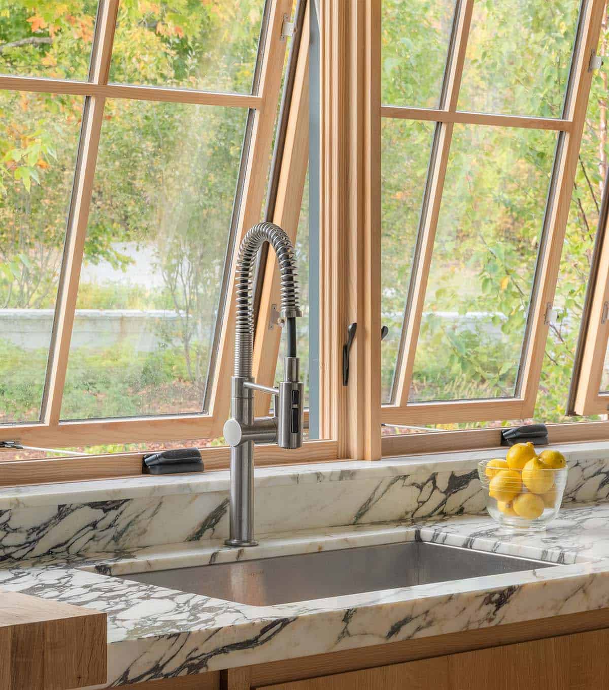 Close-up of a stainless kitchen faucet and marble sink beneath tilted awning windows