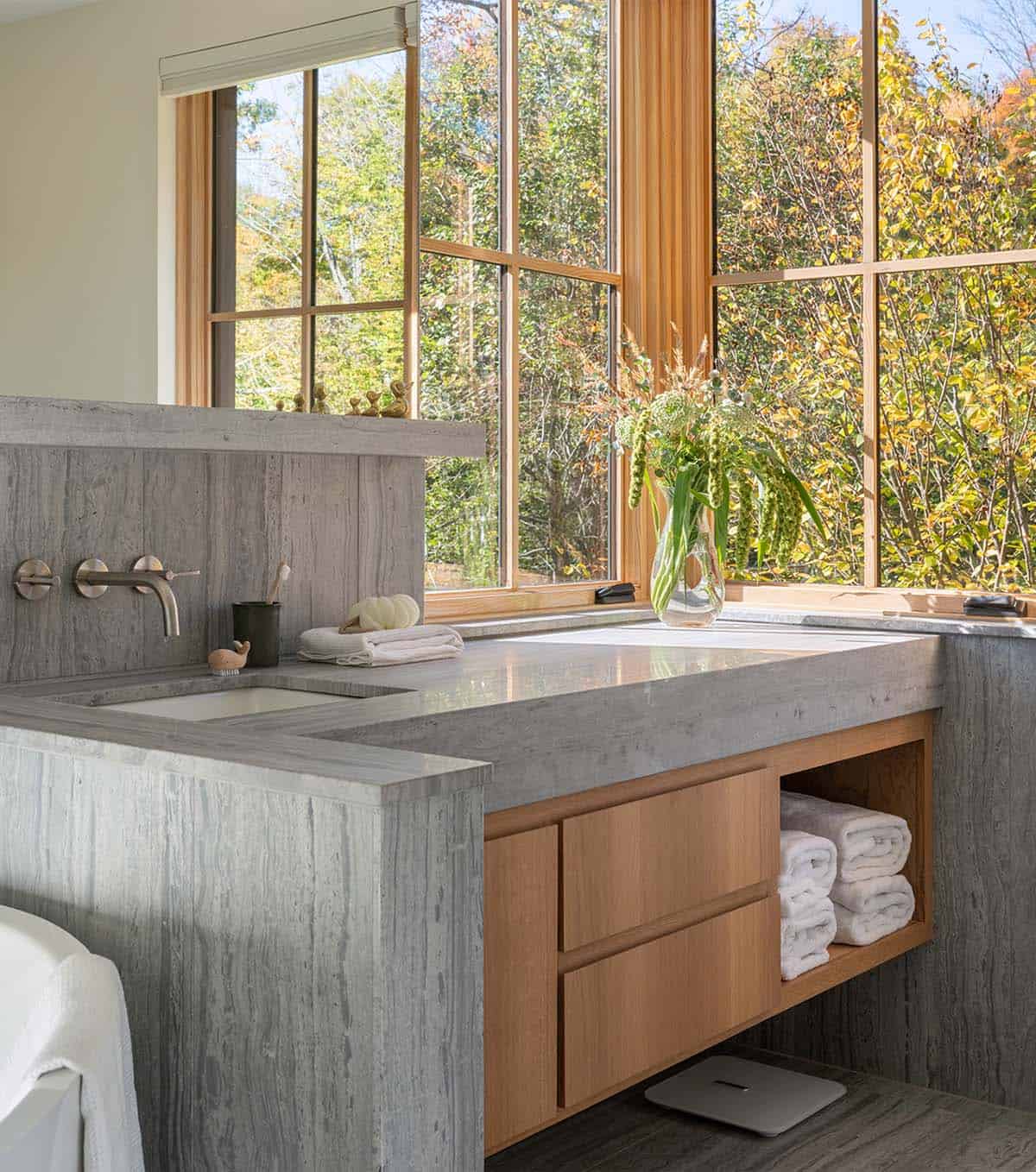 Bathroom vanity with stone countertop, wall-mounted faucets, and a vase of greenery by the window